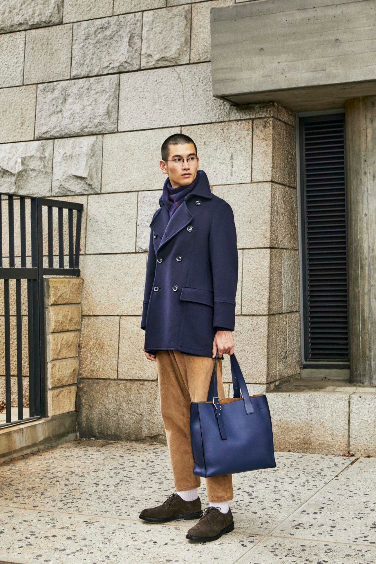 Young man in navy peacoat and brown trousers holds blue leather bag, standing against stone building wall