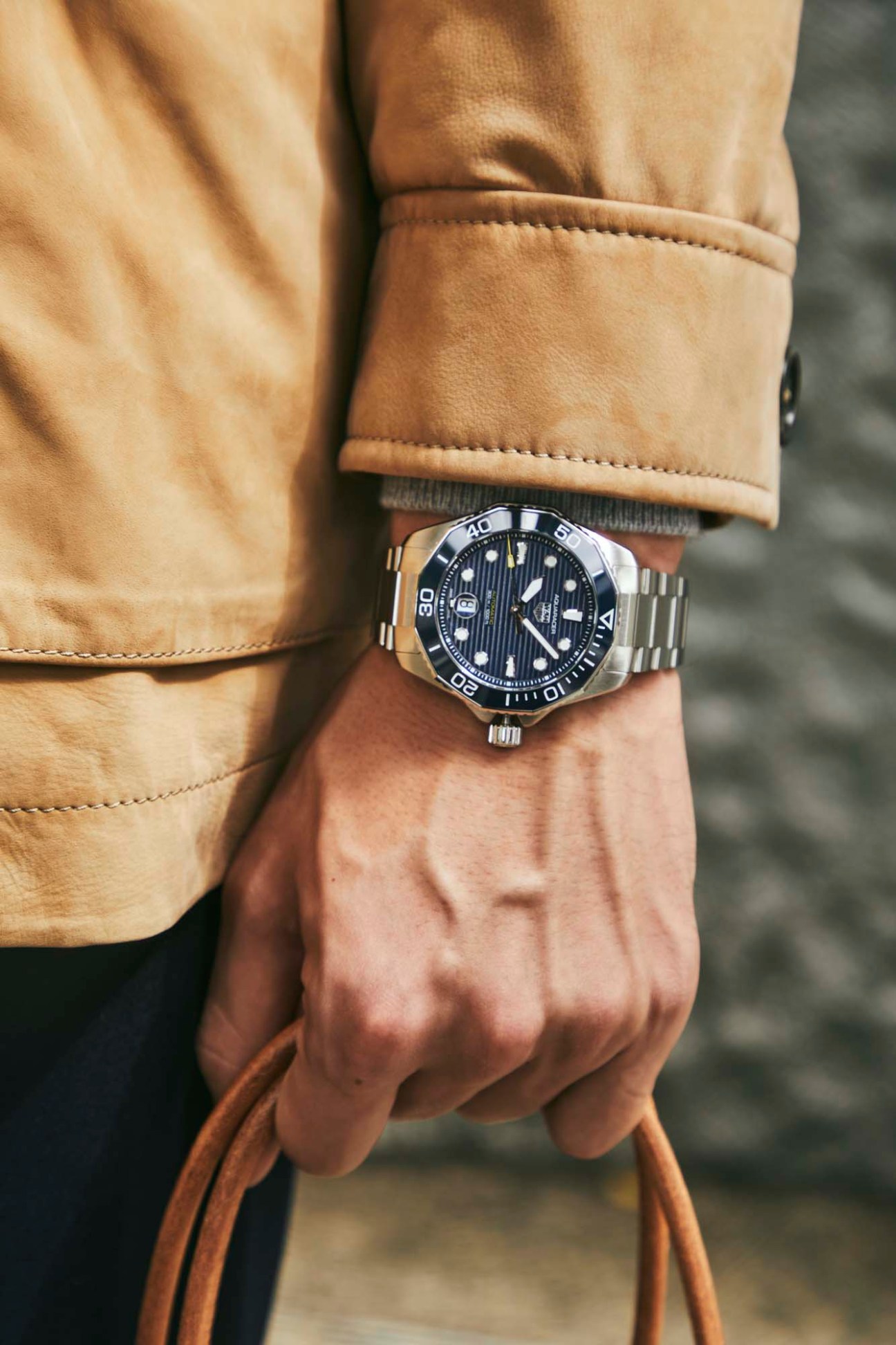 Person wearing camel-colored wool coat with detailed stitching and blue-faced diving watch on wrist