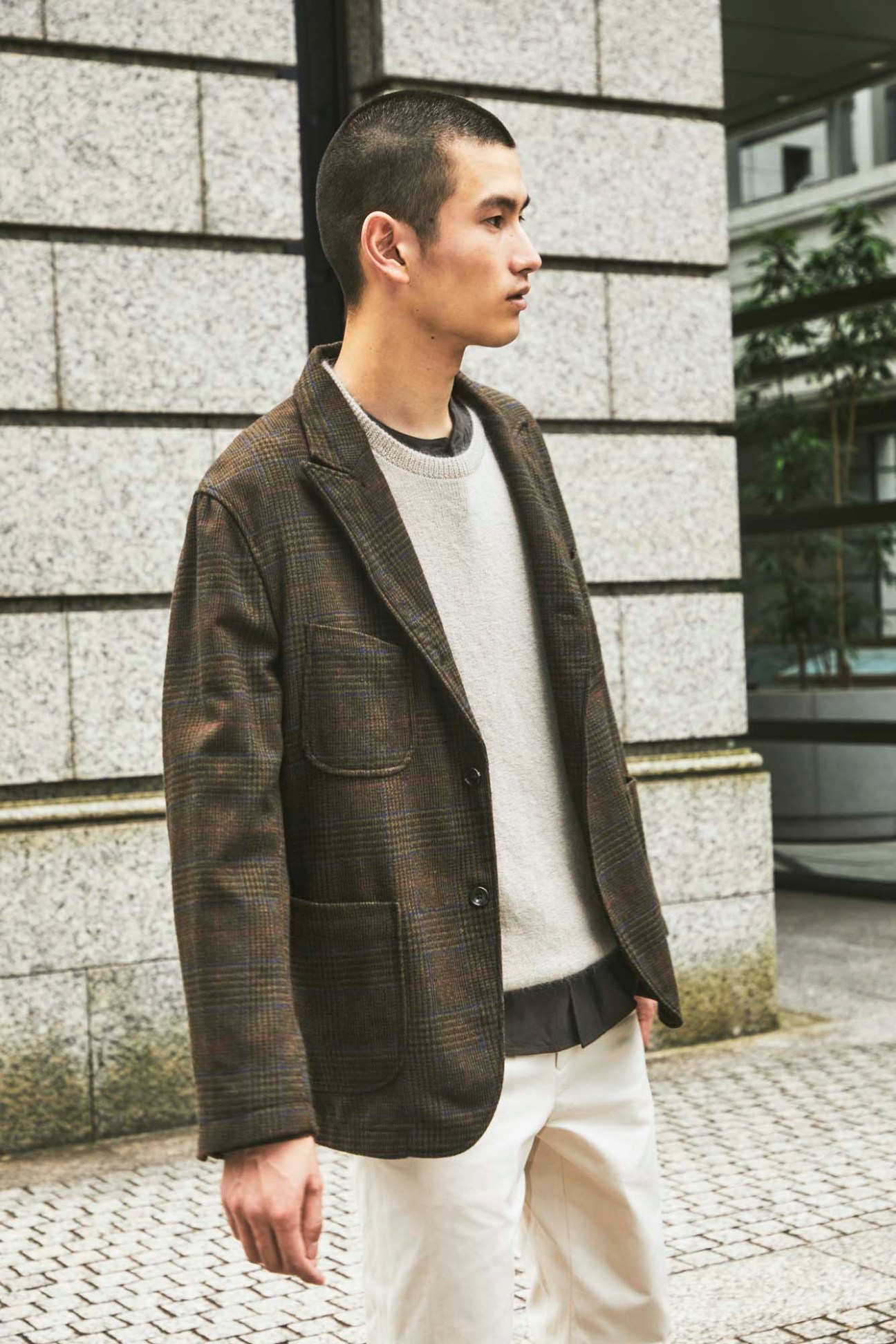 Young man in brown checked blazer and cream sweater stands on cobblestone street against concrete building