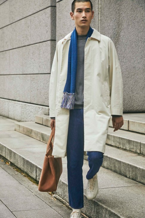 Asian man in cream trench coat, blue scarf, striped shirt and navy pants walking on concrete steps with brown leather bag