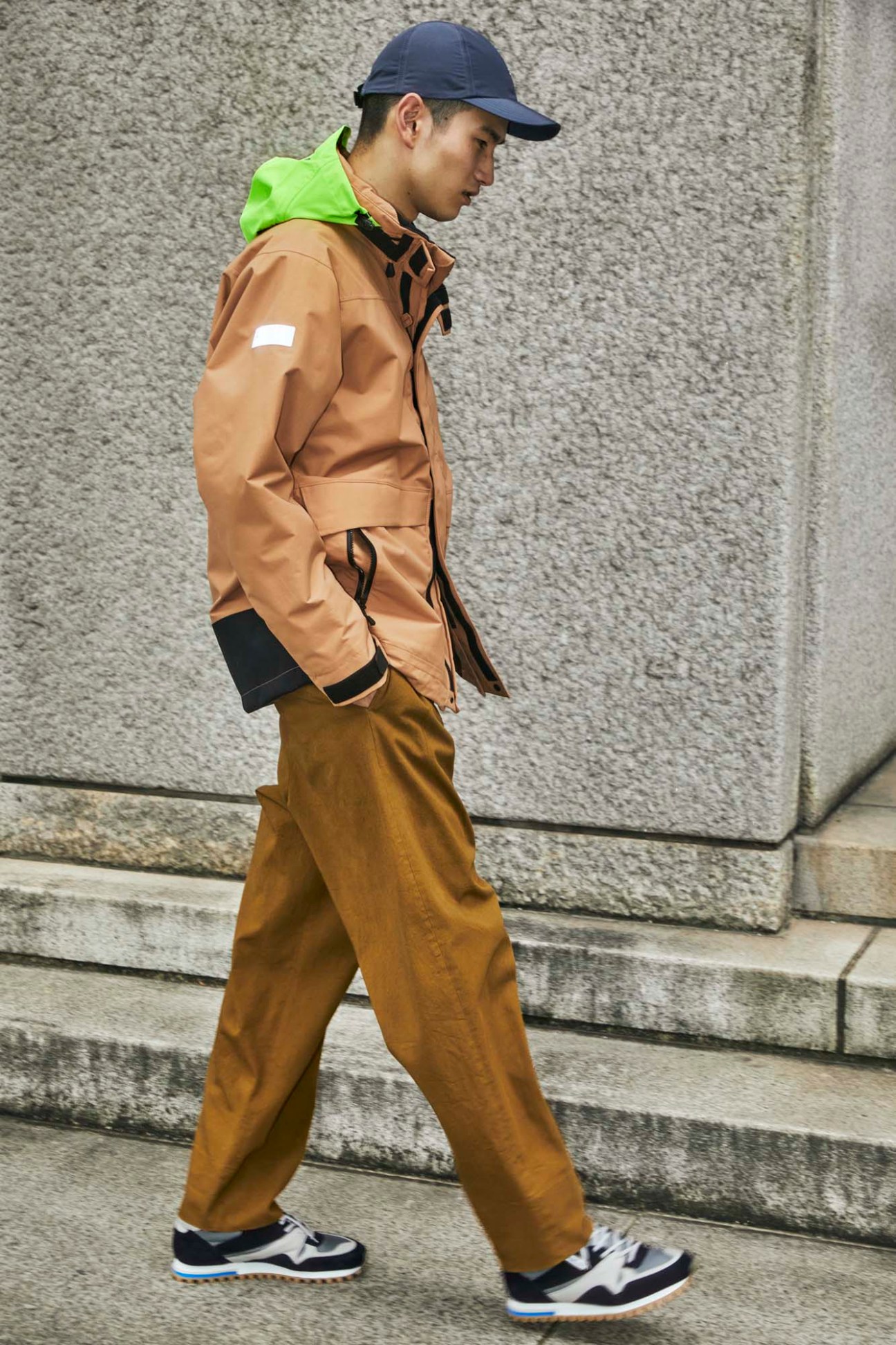 Young man walking down stone steps wearing tan jacket, brown trousers, navy cap and sneakers