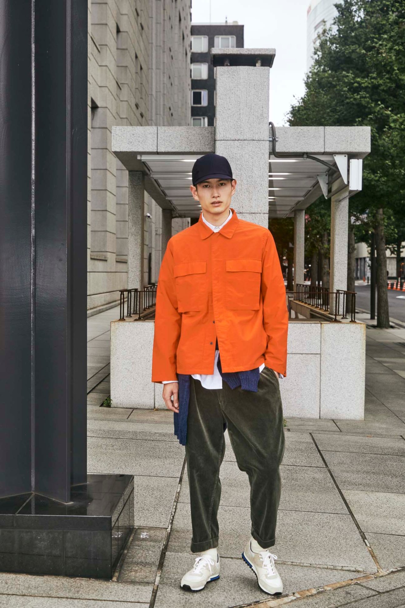 Young man in orange jacket, olive pants and navy cap standing on urban steps in modern Japanese cityscape