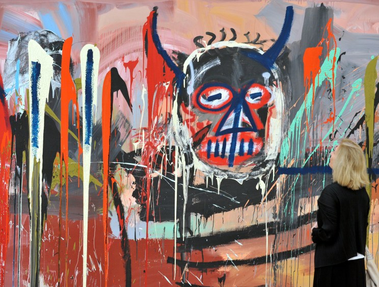 A visitor views a large-scale abstract painting with a horned skull face featuring red and blue markings in vibrant colors.