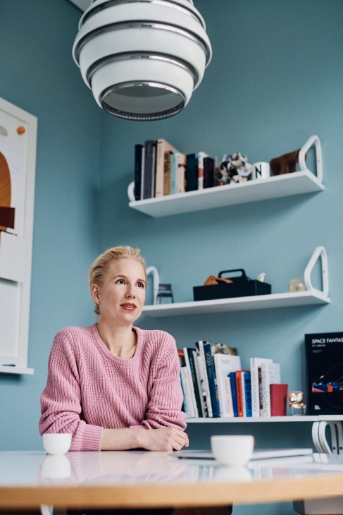 Nora Fehlbaum sits at a desk in her office near Basel, wearing a pink sweater, with blue walls and white shelving behind her.
