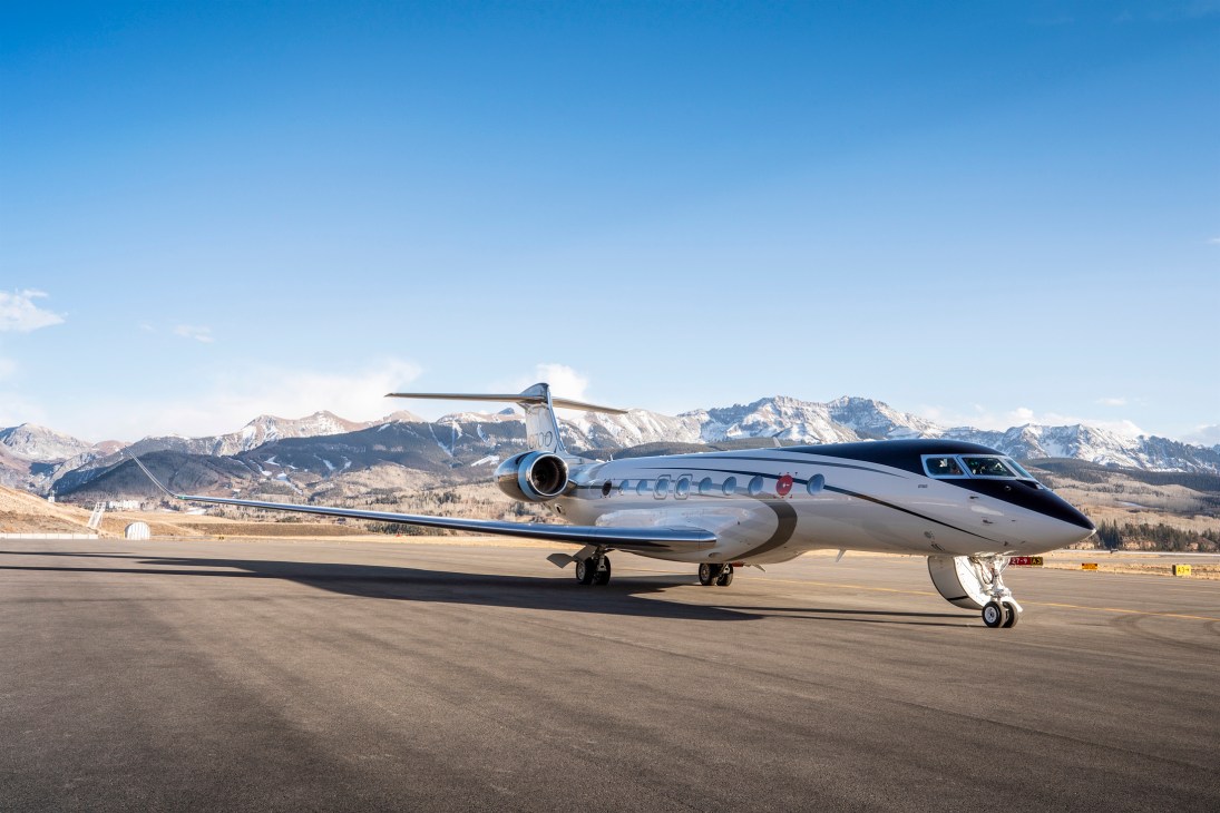 White Gulfstream G700 private jet parked on tarmac with snow-capped mountains in background under clear blue sky