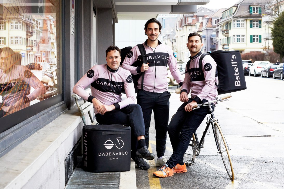 Three Dabbavelo delivery workers in pink and black uniforms posing with bicycles and delivery bags on a European street