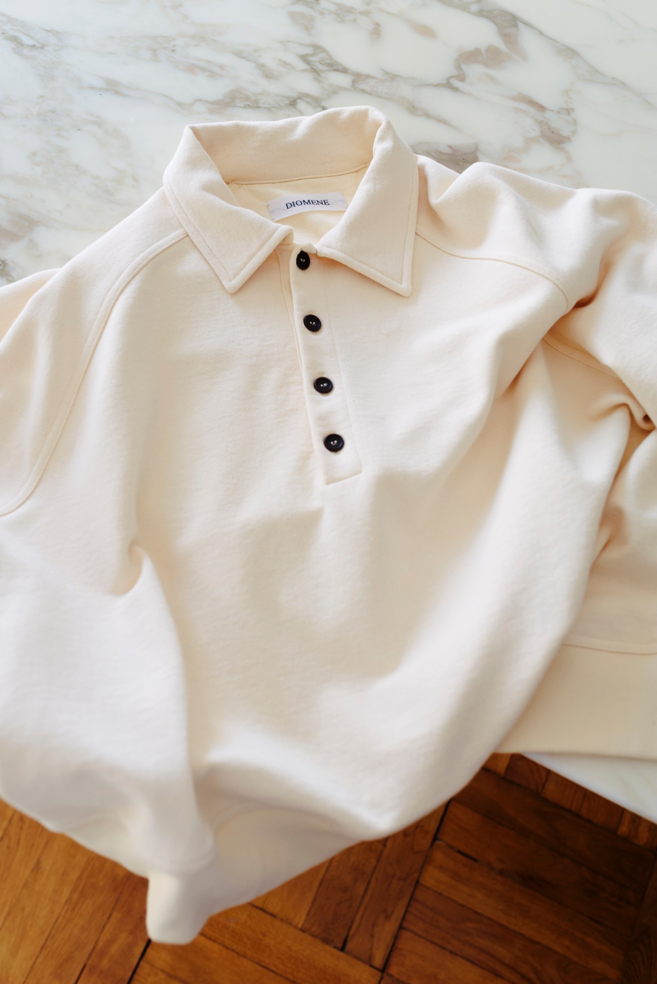 Cream-colored Diomene polo shirt with collar and black buttons laid flat on marble surface