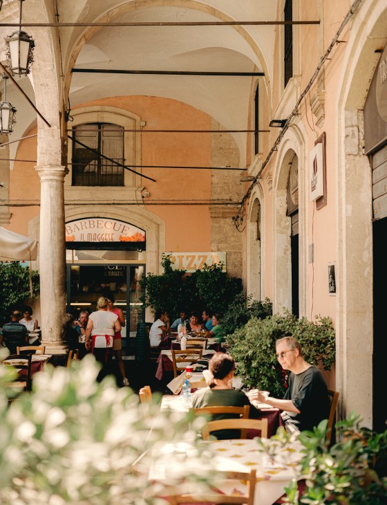 People dining at outdoor tables under an arched colonnade in a historic Catania street.