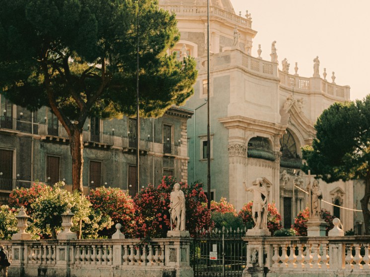 A cream-coloured baroque church with a domed cupola surrounded by flowering plants and trees in Catania's central piazza.
