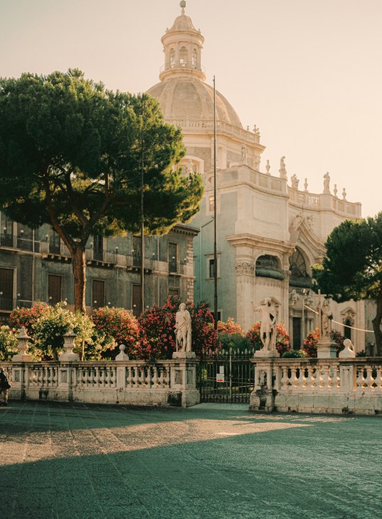 A cream-coloured baroque church with a domed cupola surrounded by flowering plants and trees in Catania's central piazza.