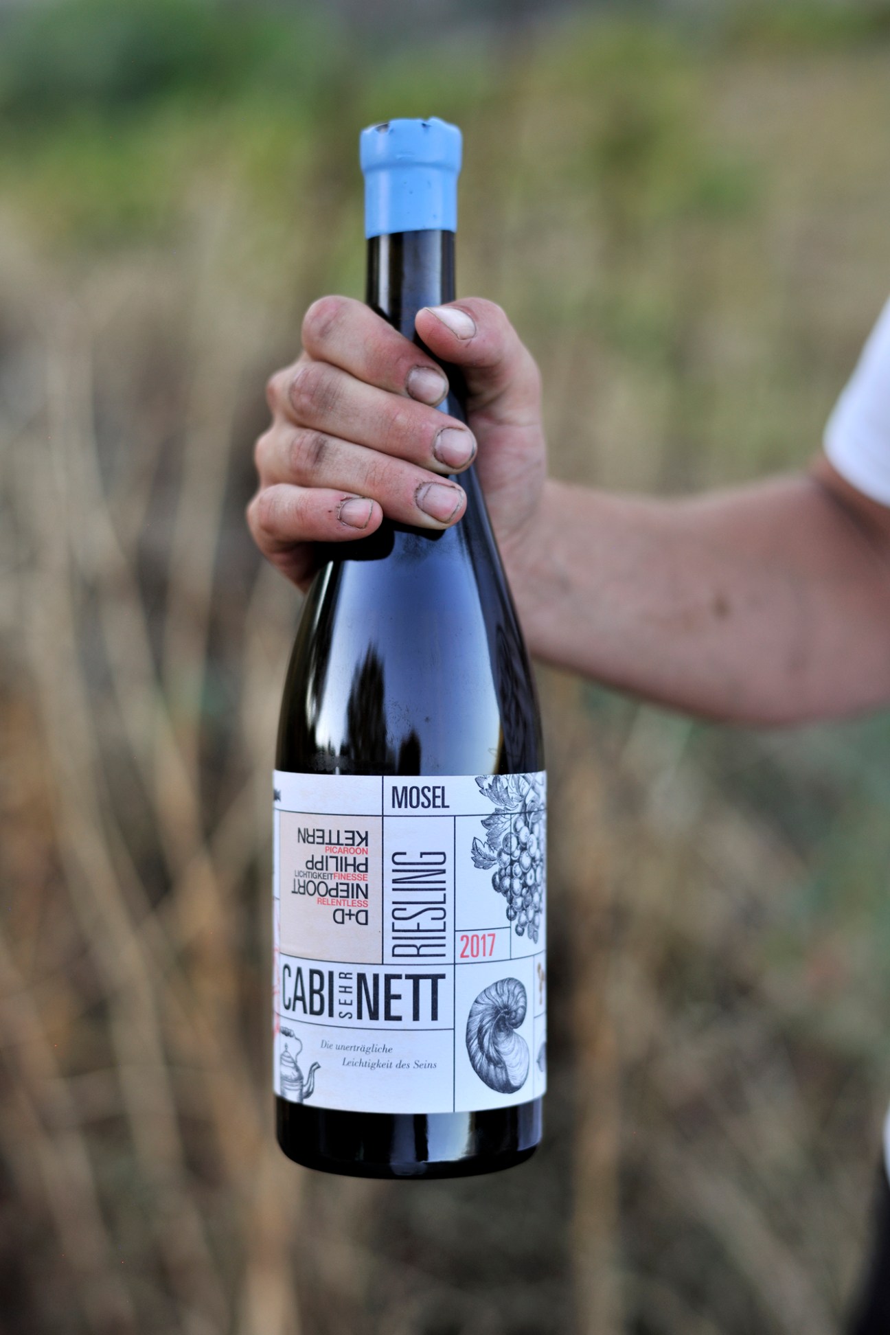 Hand holding a dark wine bottle with blue cap and white label reading Cabinet Nett, Riesling 2017, Mosel