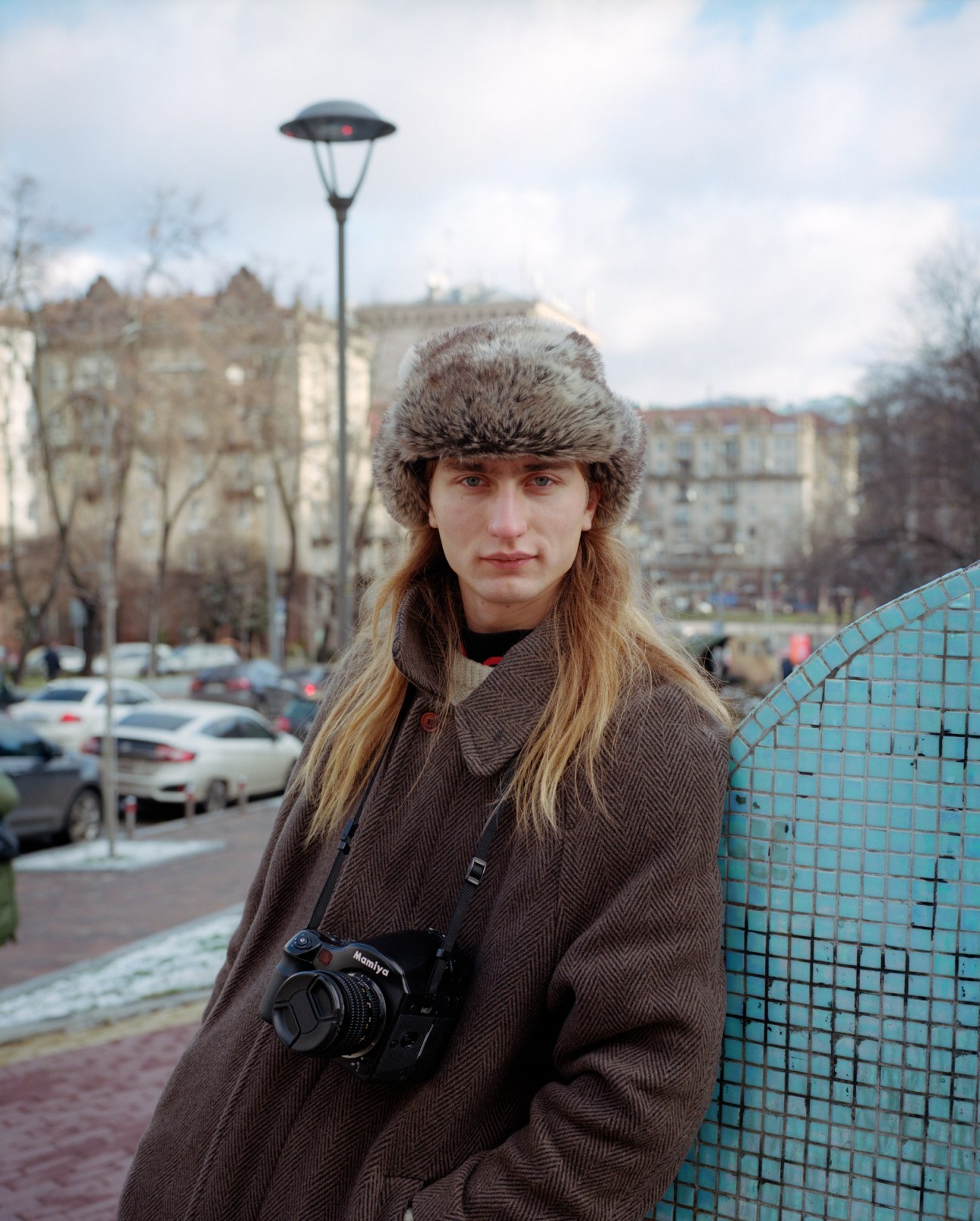 Daniil, a photographer, wearing a fur hat and brown coat with a camera around his neck in Kyiv