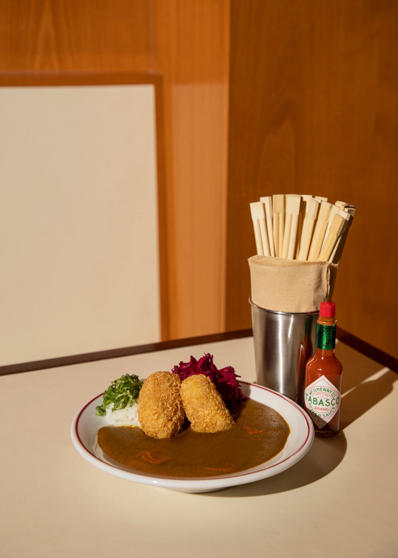 Plate of golden-fried croquettes with curry sauce, rice, and purple cabbage garnish beside chopsticks and Tabasco sauce