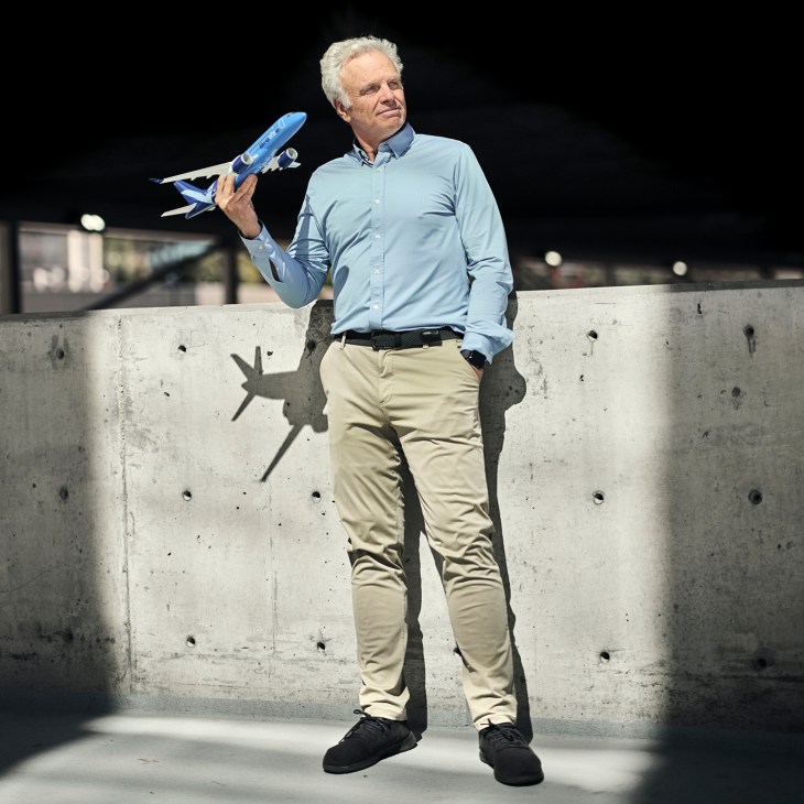 David Neeleman holds a blue model airplane while standing against a concrete wall.