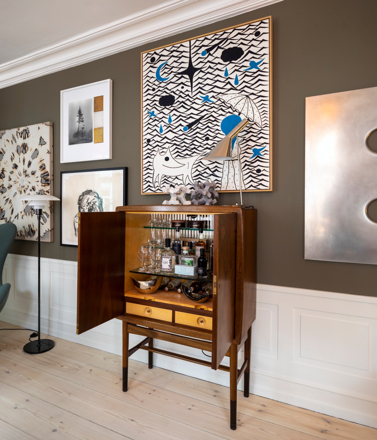 Mid-century wooden minibar cabinet with glass shelves displaying bottles and glassware, beneath contemporary artwork on taupe wall.