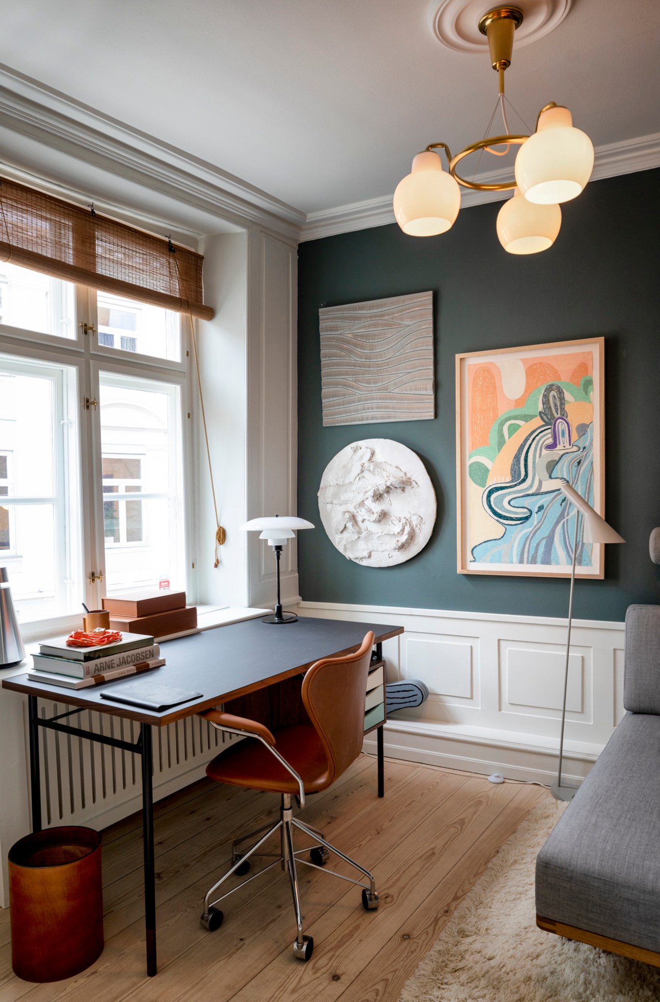 Modern office space with desk, leather chair, contemporary artwork on dark walls, and brass pendant lighting.