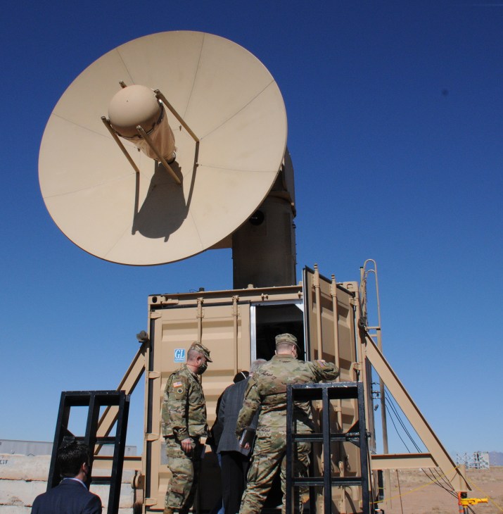 Military personnel in camouflage uniforms operating a large white satellite dish communication system