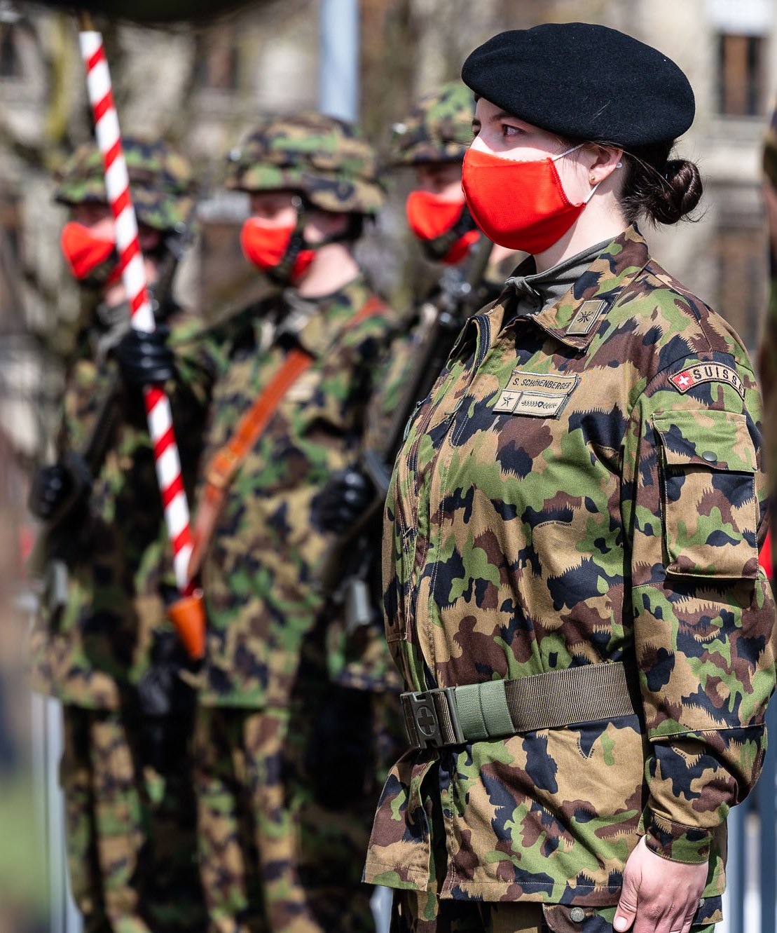 Female Swiss military officer in camouflage uniform and red face mask standing at attention during ceremony