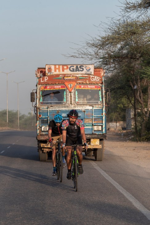 Two cyclists in helmets and athletic gear riding high-end bikes on a New Delhi road ahead of a colorful painted truck.