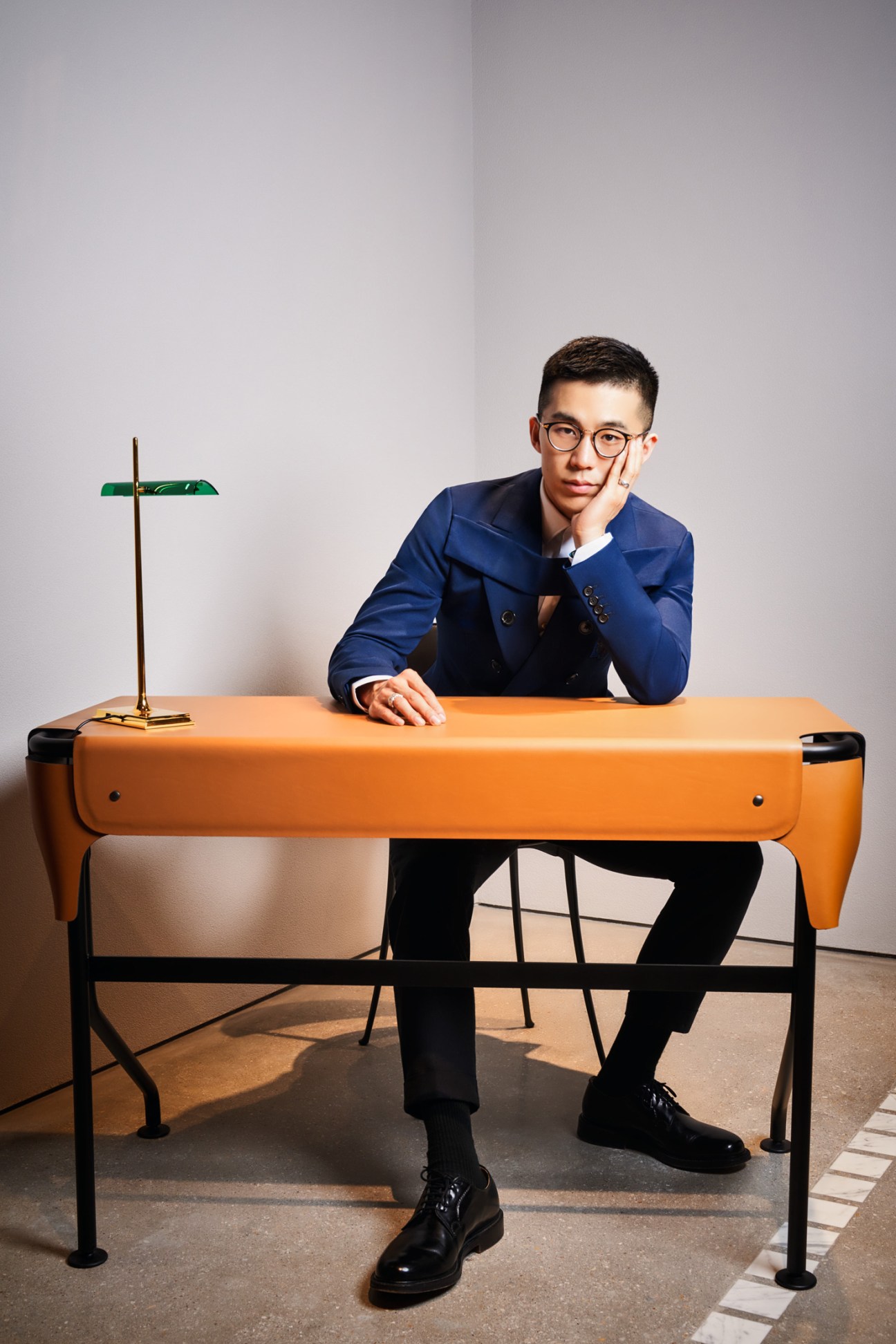 Demos Zhu, founder and CEO of Mingus furniture, seated at an orange desk wearing glasses and a blue blazer.