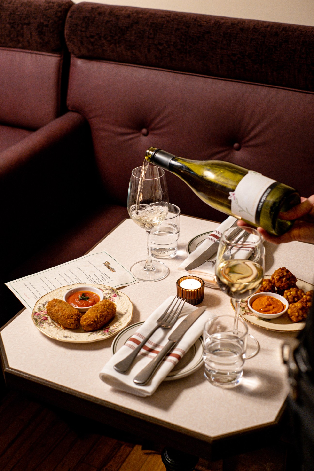 Hand pouring white wine into glass on restaurant table with chicken croquettes and elegant table setting