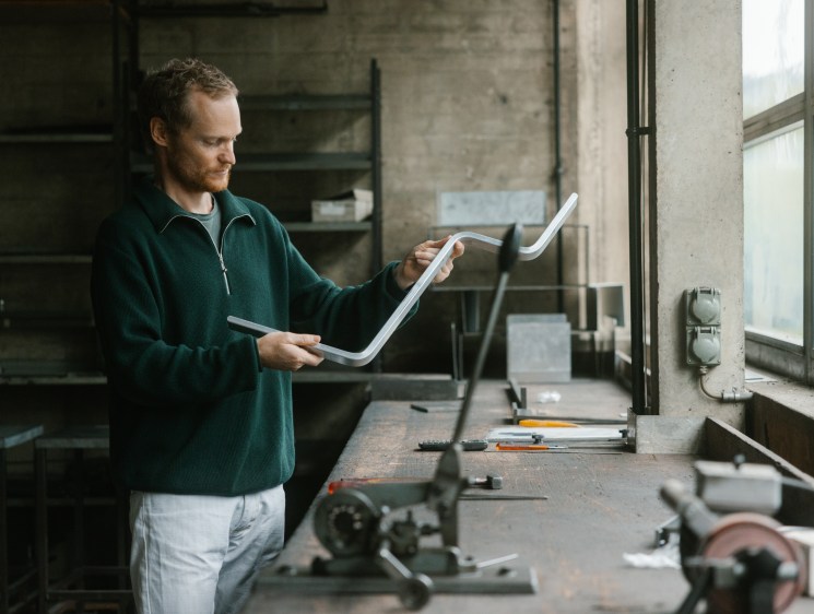 Man in green sweater examining white metal chair component in workshop with machinery