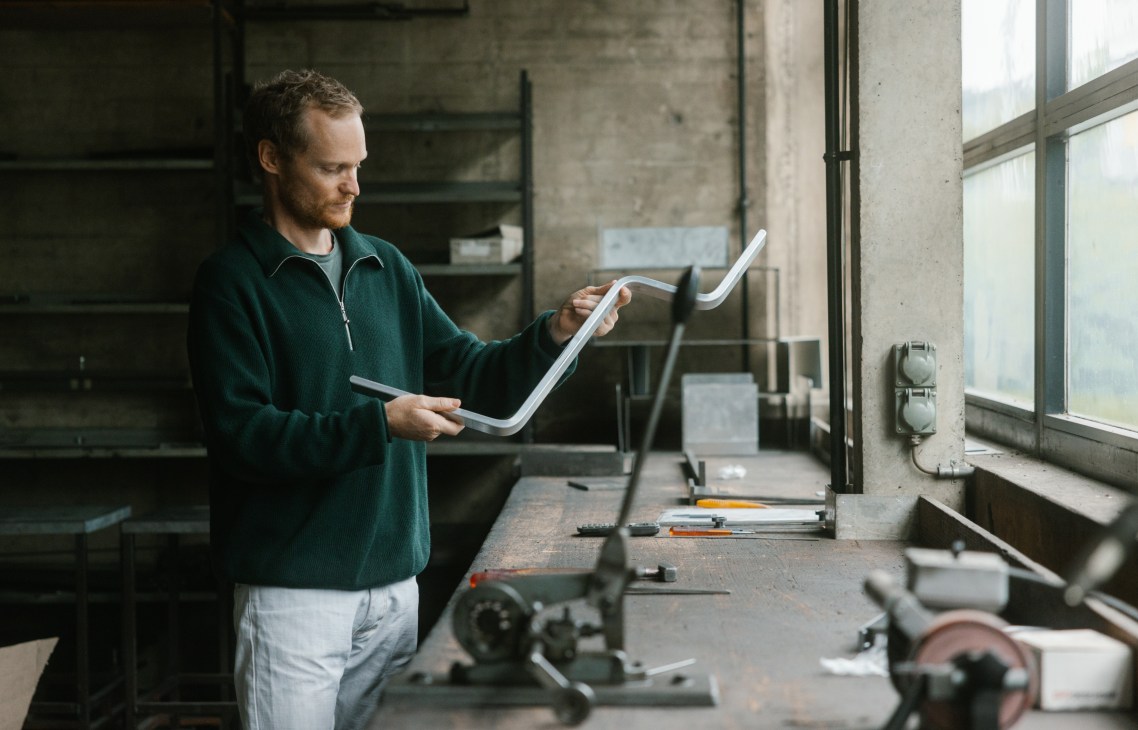Man in green sweater examining white metal chair component in workshop with machinery