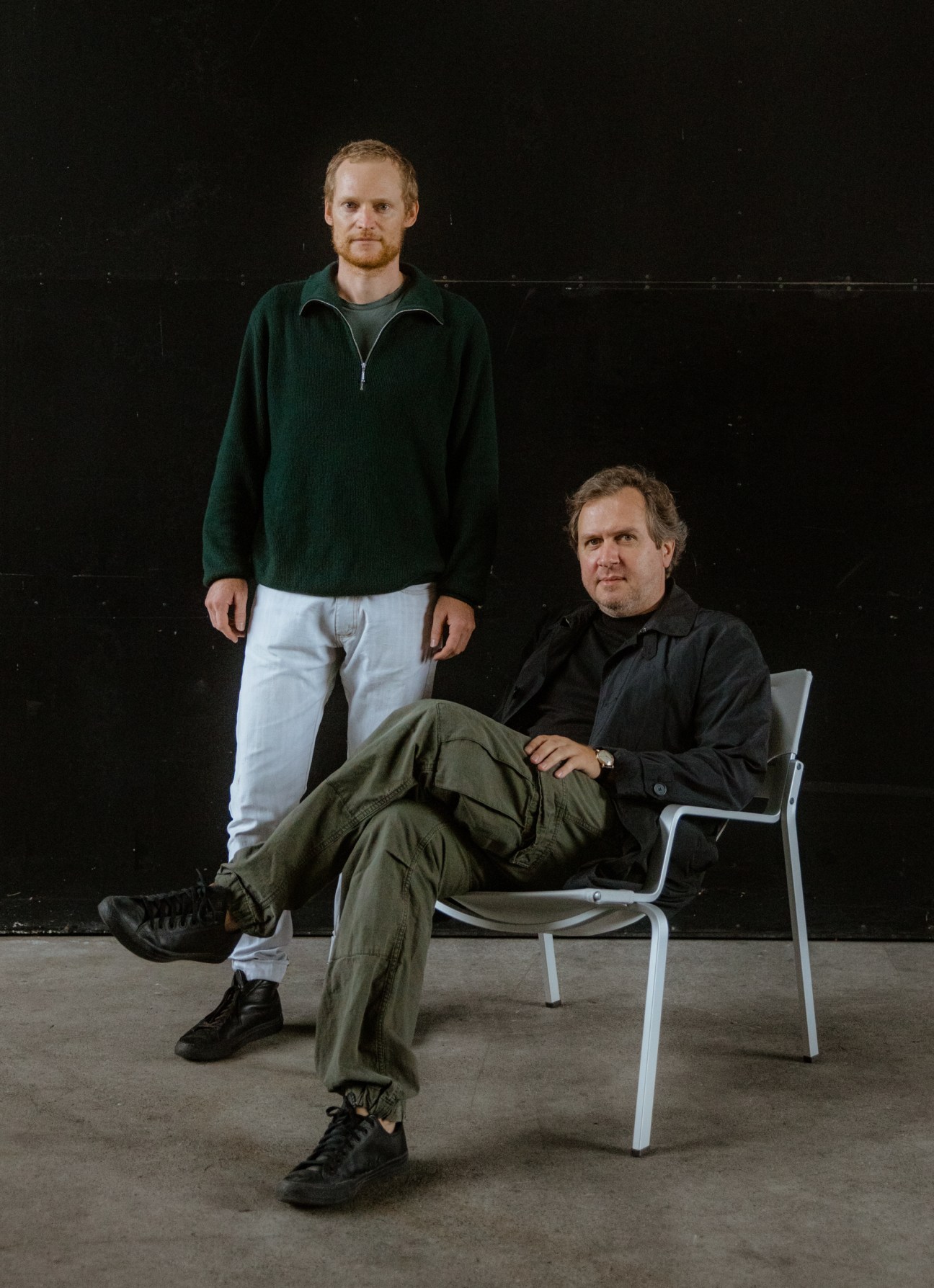 Two men with one standing wearing green pullover and one sitting on aluminum chair in dark industrial setting