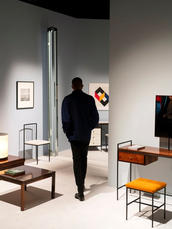 Person in dark clothing viewing furniture display with chair, desk, and artwork in gallery booth
