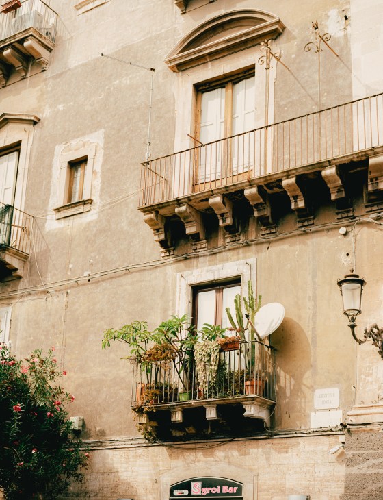 Ornate Catania building facade with decorative balcony and potted plants, showing volcanic stone architecture.