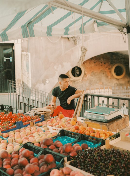 Young vendor at the Pescheria market in Catania selling fresh produce under a striped awning.
