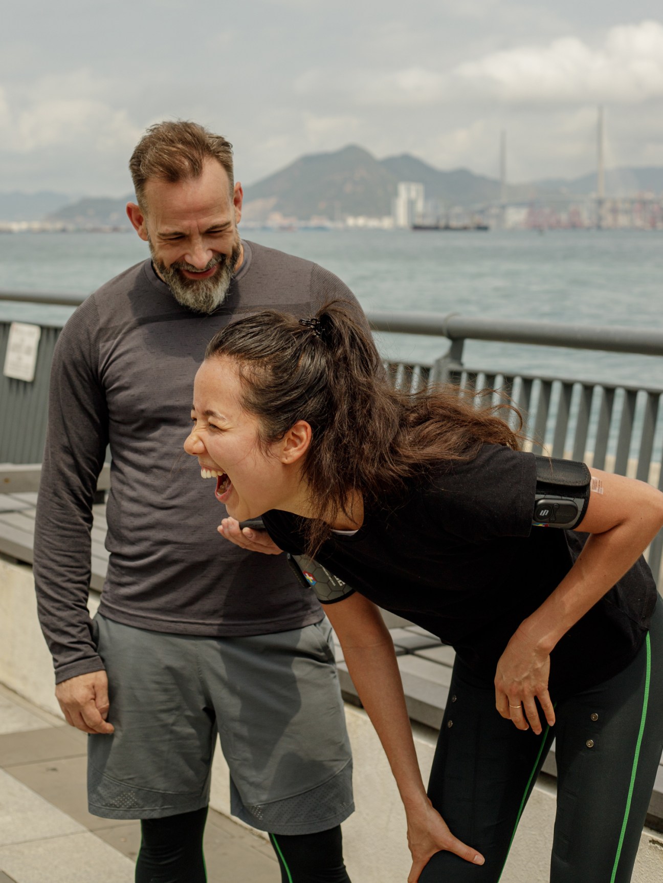 Man and woman in athletic wear exercising outdoors by water with city skyline in background.