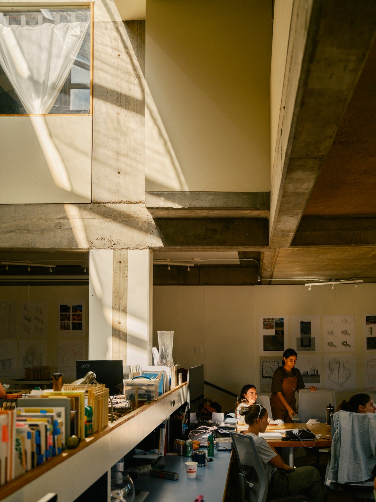 Architecture students work at desks in a studio space with concrete ceiling and natural light streaming through windows.