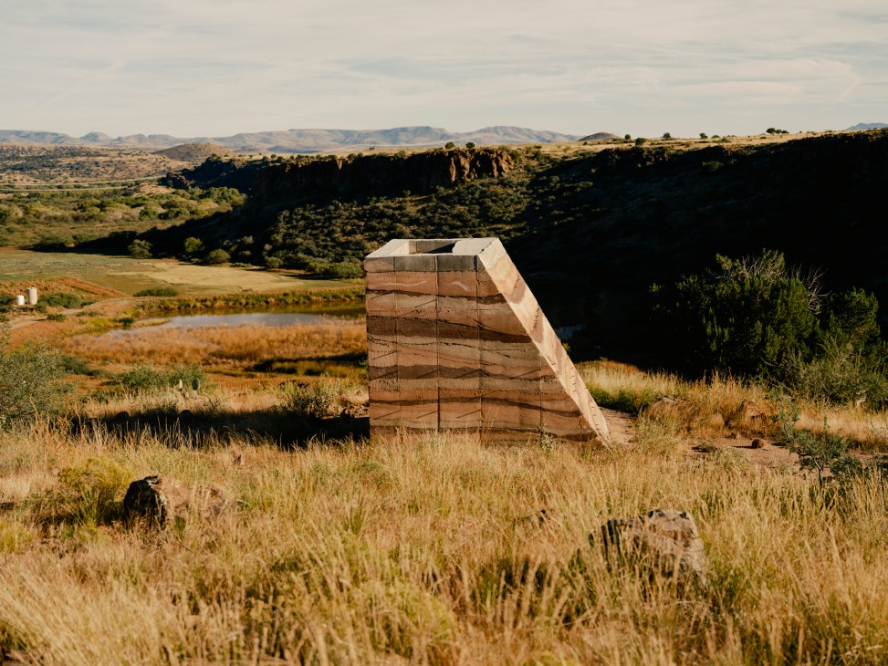 Jessica Martin's rammed-earth trapezoid shelter 'Cinders' in a desert landscape with canyon and grassland.