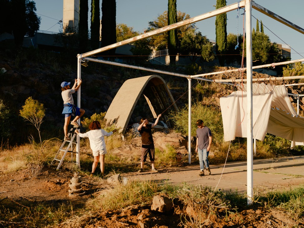 Architecture students constructing a temporary shelter frame with white metal pipes and canvas in an Arizona desert setting.