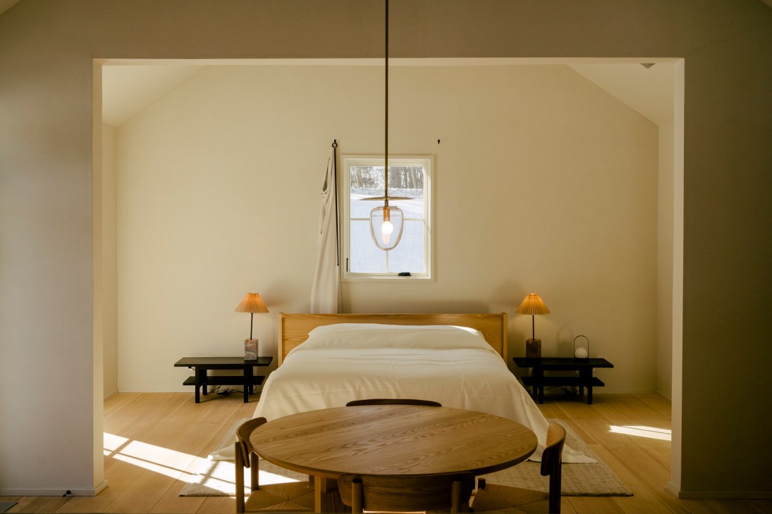 Minimalist hotel room with wooden bed, white bedding, dark side tables with lamps, and round wooden table
