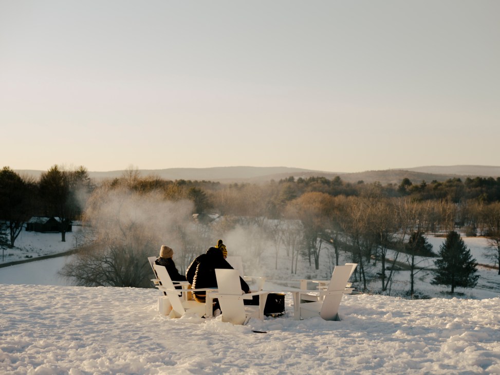 Two people sitting in white outdoor furniture around a fire in snowy landscape with rolling hills