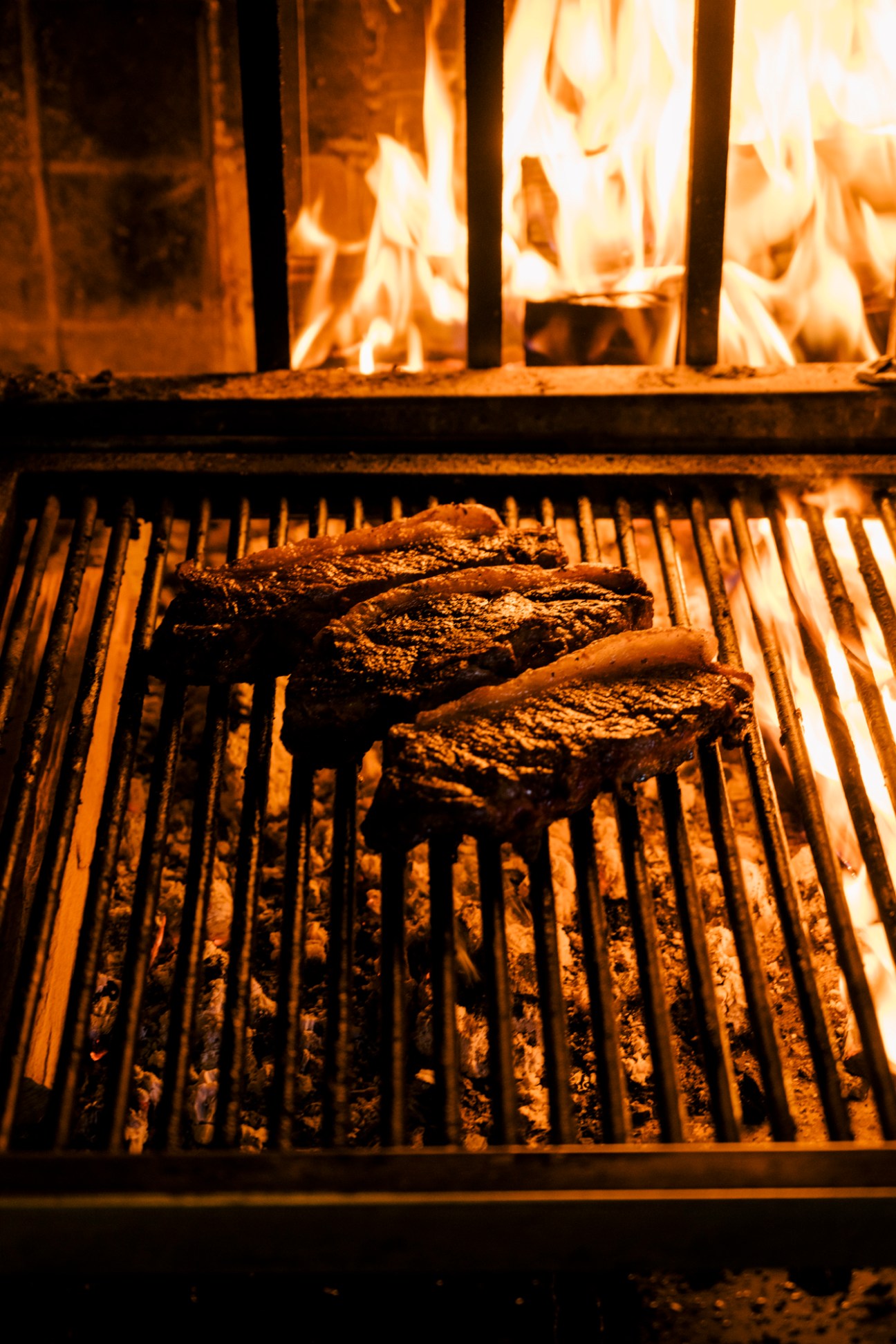Steaks cooking on metal grill grates over open flames in fireplace