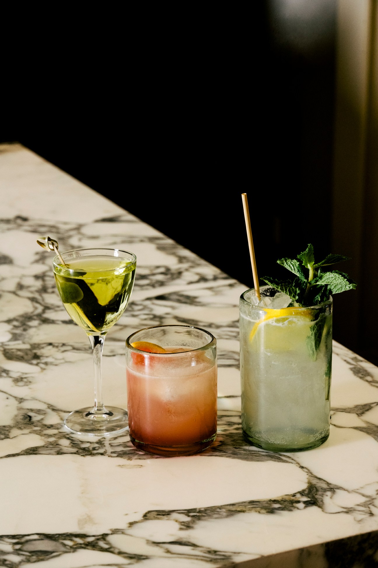 Three cocktails on a marble table - a green martini, an orange-pink drink, and a pale drink with mint and gold straw