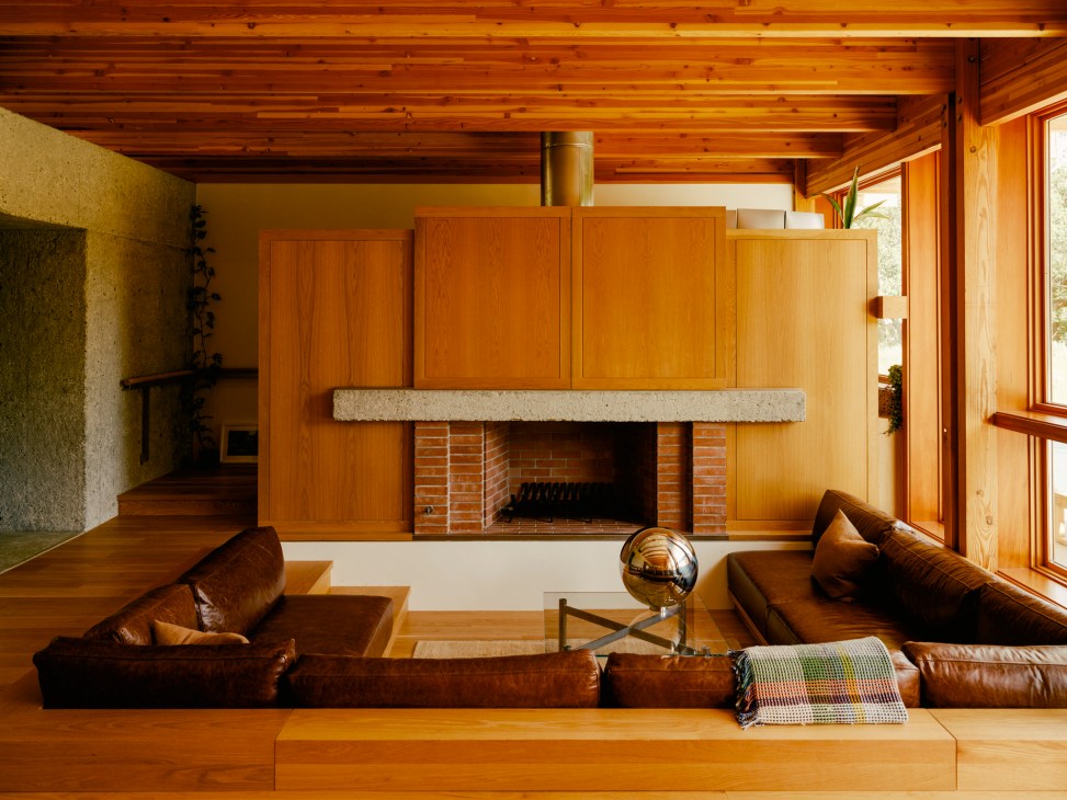 Sunken living room with built-in leather seating, exposed wood ceiling beams, and modernist fireplace with brick surround