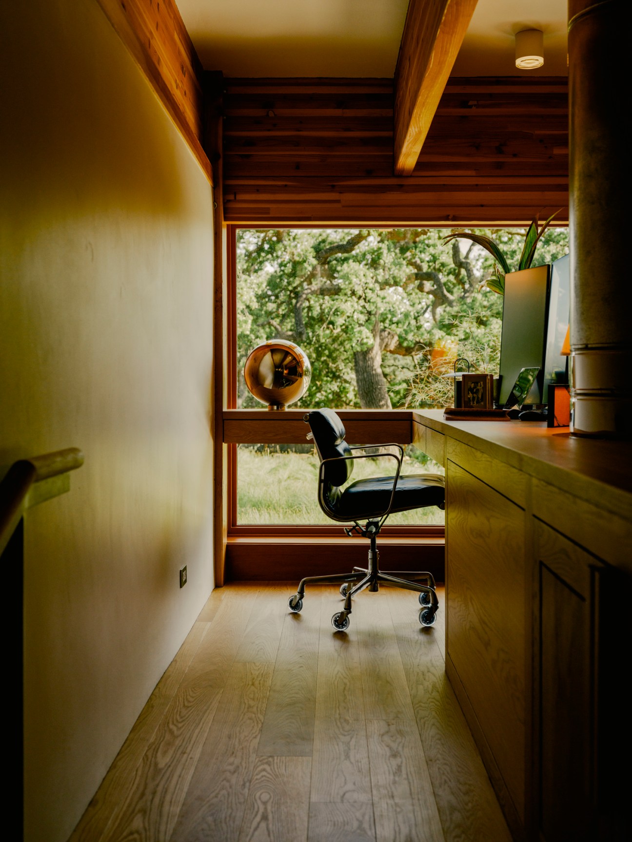 Modern loft office with exposed wood beams, built-in desk, black office chair and large window overlooking trees