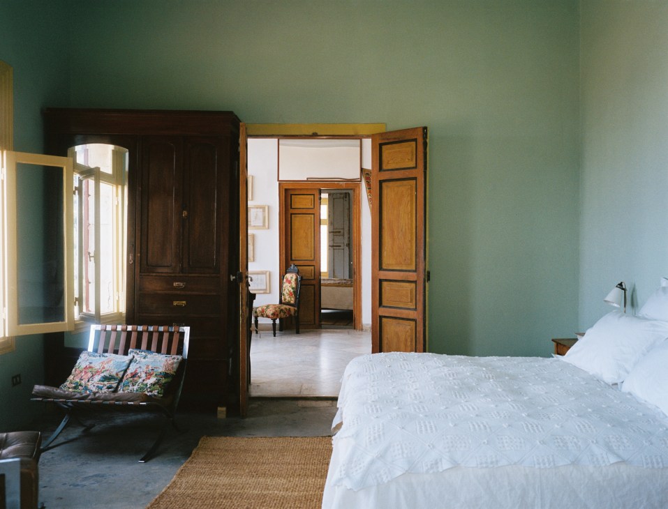 Bedroom with green walls, dark wooden wardrobe, striped bench, white bedding and view through doorway to adjacent room