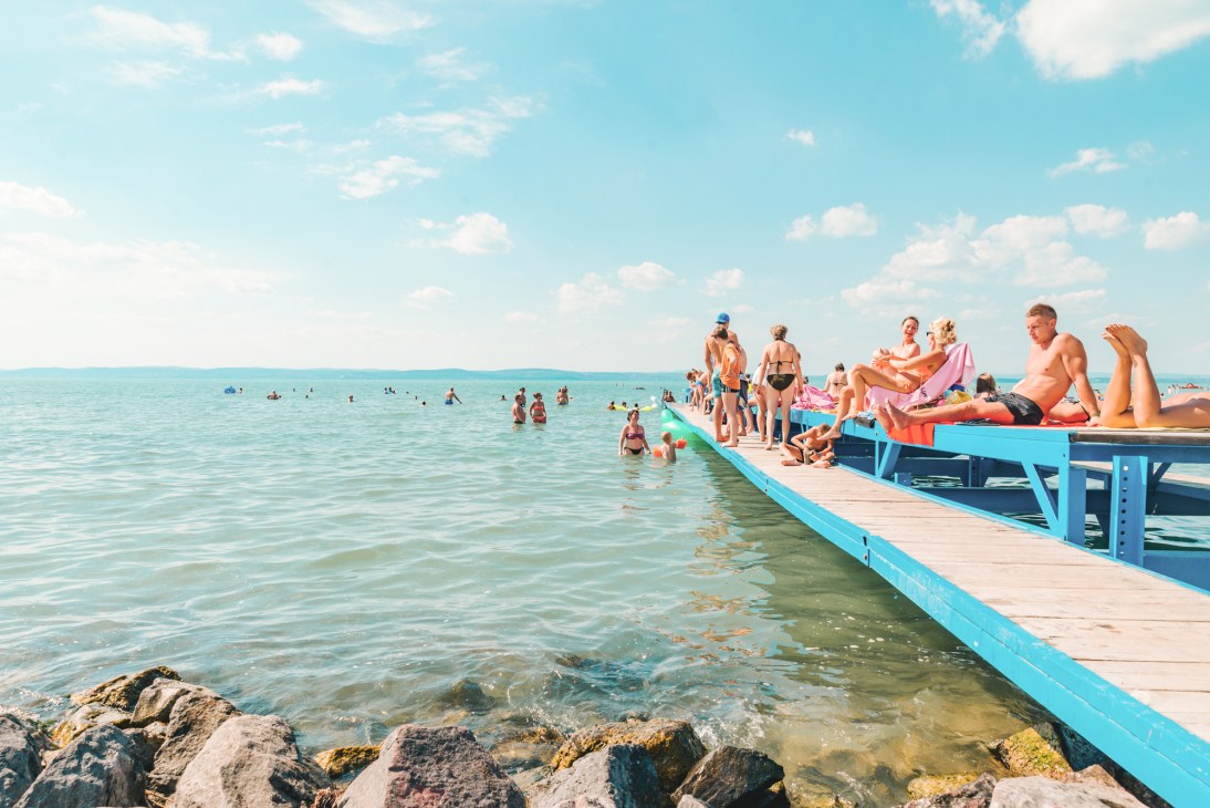 People sunbathing and swimming on a wooden pier extending into Lake Balaton with clear blue water