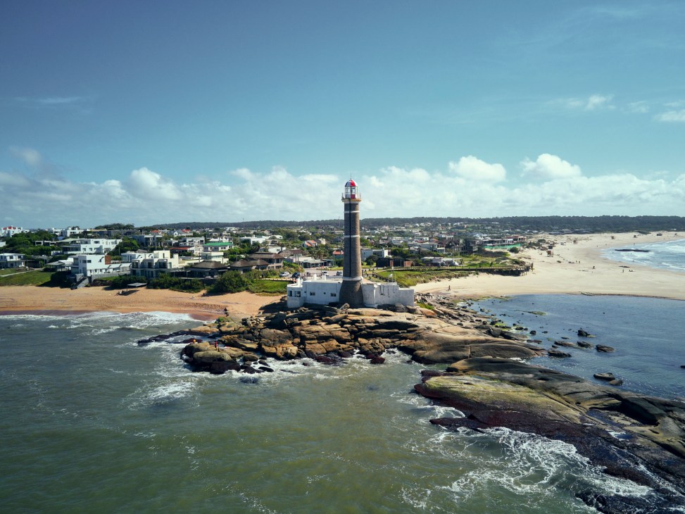 José Ignacio lighthouse with red and white tower on rocky outcrop dividing two beaches, with coastal town and homes in background.