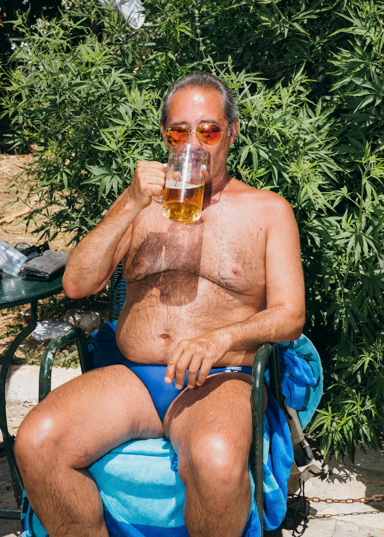 Shirtless man with sunglasses drinking beer from glass mug while sitting in blue chair outdoors