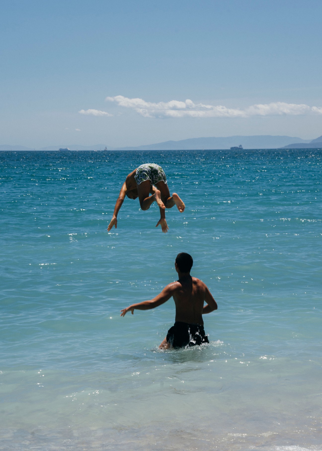 Person diving into blue ocean water while another person stands waist-deep in the sea