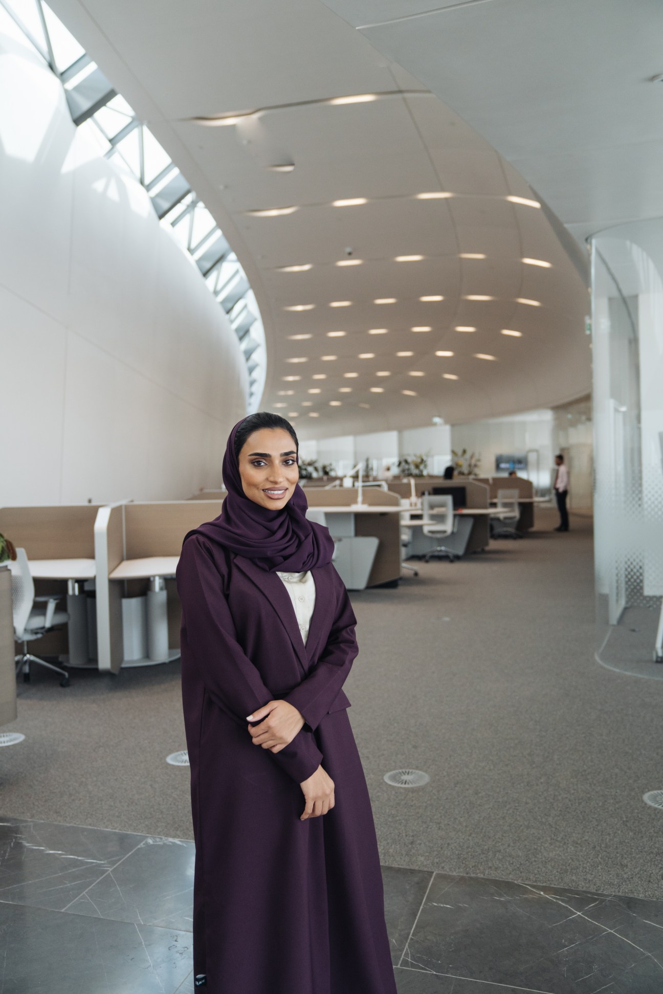 Protocol officer Noura Al Shara standing in a modern office corridor with curved ceiling and recessed lighting at Beeah HQ.