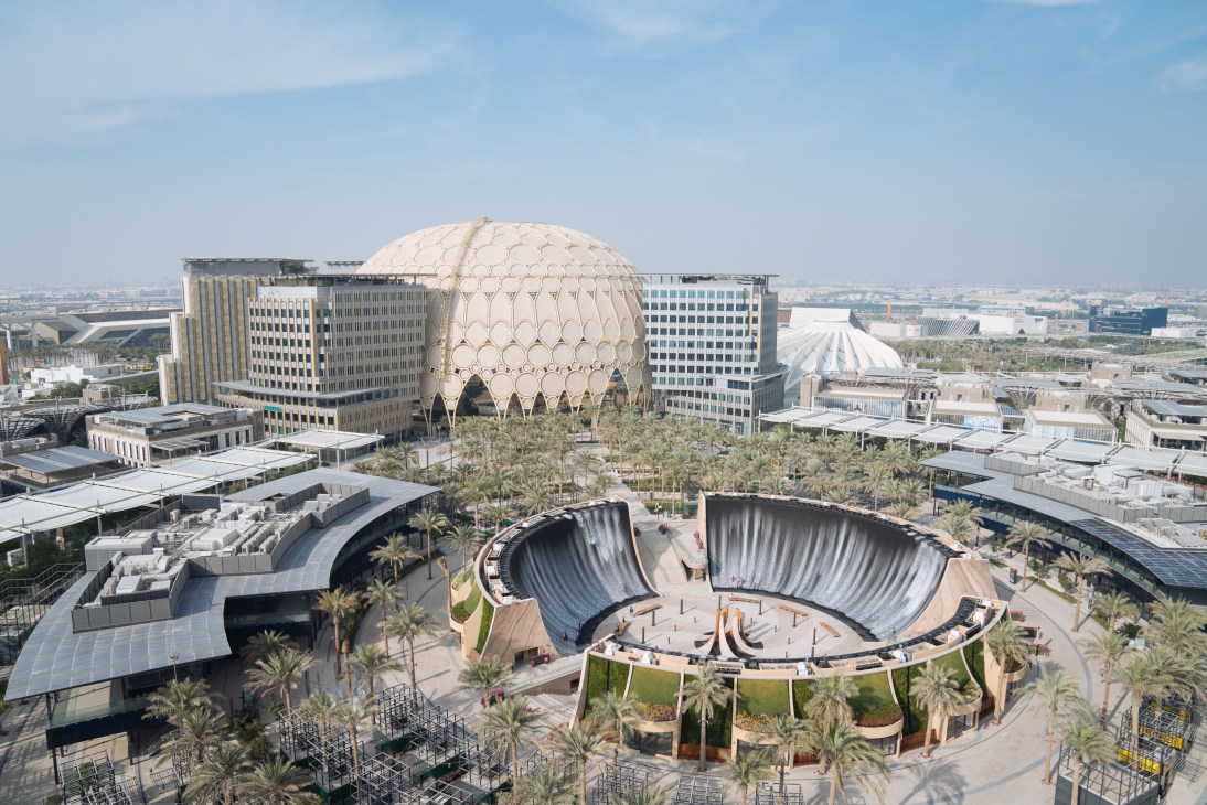 Aerial view of Expo City Dubai showing the distinctive cream-colored domed Al Wasl Plaza surrounded by modern office buildings and palm trees.
