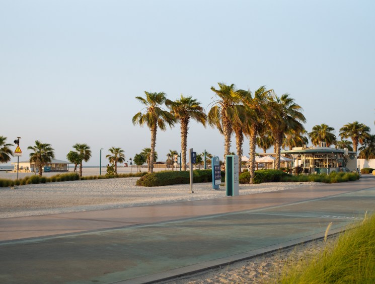 Palm-lined jogging track along Jumeirah Beach with sandy shore and cafés in the distance.