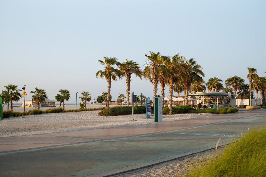 Palm-lined jogging track along Jumeirah Beach with sandy shore and cafés in the distance.